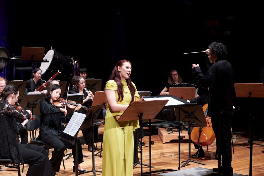 Eine S?ngerin in einem gelben Kleid steht vor einem kleinen Orchester und singt
