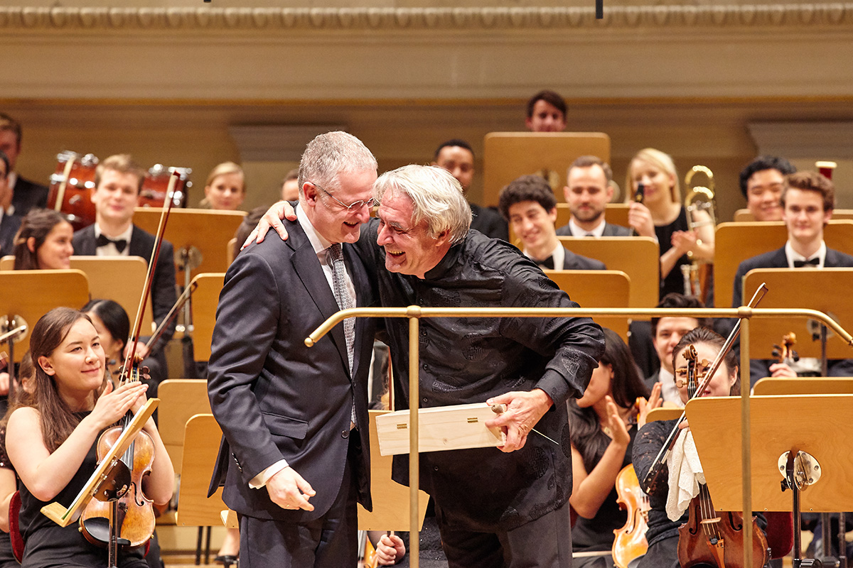 Rektor Robert Ehrlich und Professor Christian Ewald umarmen sich auf der Bhne vor dem Orchester aus Studierenden